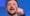 A man with a beard is speaking passionately, pointing his finger as if addressing an audience about corruption. He stands in front of a blue background, reminiscent of an Ethicswatchnj broadcast, adding a sense of gravity and purpose to his words.