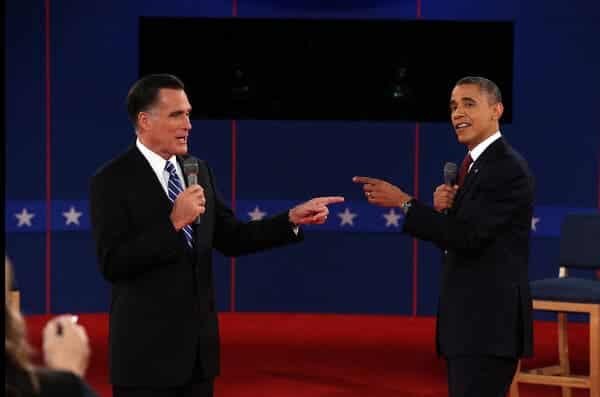 Mitt Romney and Barack Obama on a stage, both holding microphones and pointing to each other,