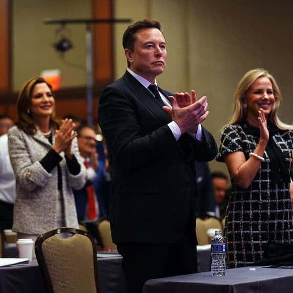 Elon Musk claps while standing in a conference room among other people who are also standing and clapping.