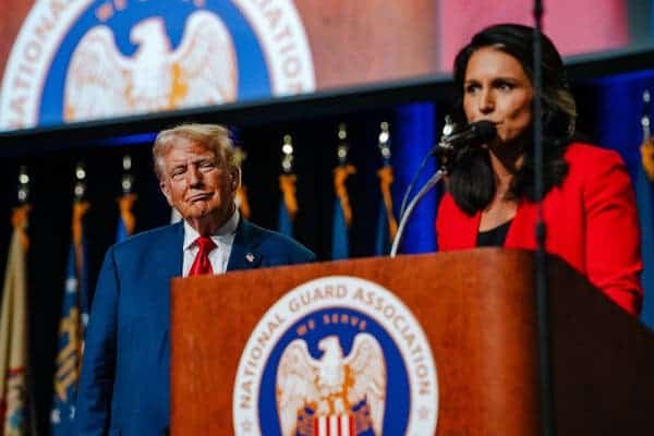 Tulsi Gabbard, wearing a red jacket over a black top, speaks from behind a lectern carrying the logo for the National Guard Association, while Donald J. Trump, in a blue jacket, a white shirt and a red tie, looks on.